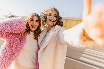 Winsome woman in pink coat having fun with friend. Blithesome lady in white attire making selfie with sister.