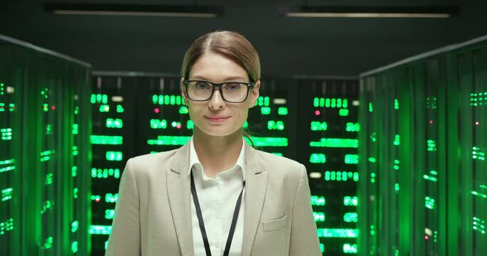 Portrait of Caucasian beautiful young woman in glasses among servers with big data smiling to camera. Green working processors in database storage. Pretty female worker of digital telecommunication.