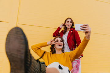 Fototapeta premium Relaxed young woman in yellow sweater making selfie with friend. Outdoor photo of two funny sisters.