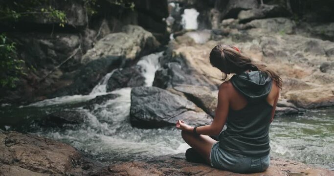 Thailand's waterfall: woman meditation pose padmasana on river bank. Relaxing leisure of girl in serenity scene of National Park at Koh Phangan Island. Cinematic footage shot in 4K, UHD