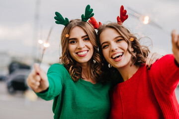 Women celebrating winter holidays. Outdoor photo of two girls with sparklers laughing at camera.
