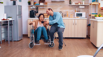 Cheerful disabled woman in wheelchair taking a selfie with husband in kitchen. Hopeful husband with...