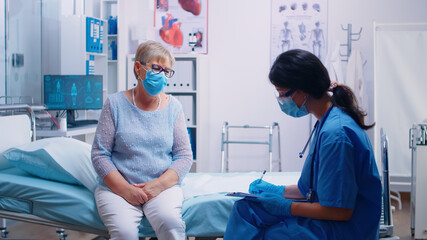 Female nurse talking with old senior patient, writing down answears. COVID 19 pandemic medical consultation. Modern private clinic or hospital. Practitioner physician appointment