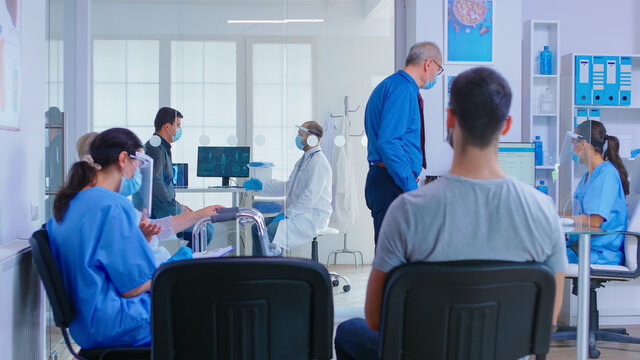 Doctor With Visor Against Coronavirus And Patient In Examination Room. Senior Man With Face Mask Talking With Nurse At Reception. Assistant And Disabled Woman With Walking Frame In Waiting Area.