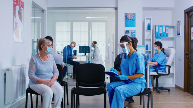 Nurse And Senior Patient Going In Hospital Examination Room From Waiting Area. Doctor Inviting Old Woman For Consultation In Office. Wearing Mask Against Coronavirus.