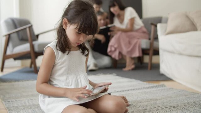 Focused young girl sitting on floor in living room and using smartphone for browsing internet while her parents and brother using tablet in background. Communication or internet concept