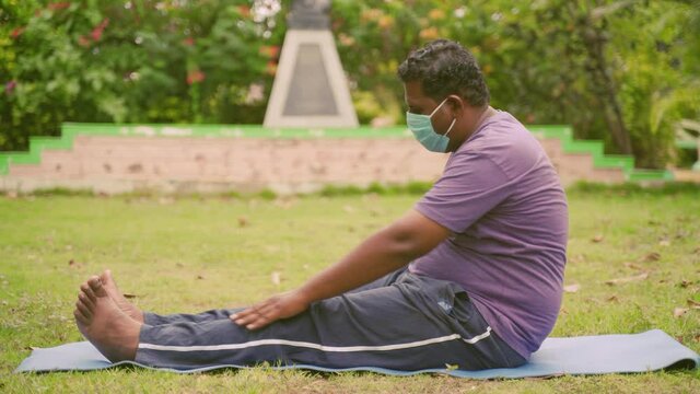 Young Fat man in medical mask exercising by trying to touch legs on yoga mat at park - Concept of Healthcare, fitness and lifestyle.