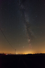 Milky Way on a summer night in Poland. A village is visible on the horizon. Poles with cables break the skyline. A car is passing nearby, leaving the white-red streaks behind.
