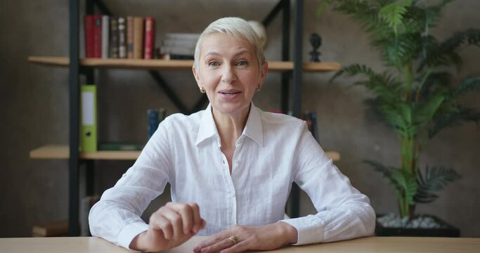 Confident elderly woman teacher in white shirt making video call, online talking to webcam at classroom