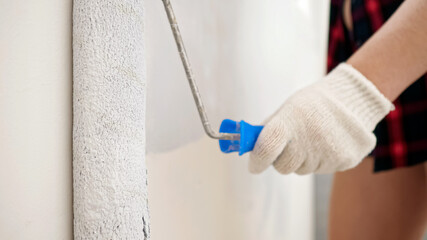 young woman in white gloves paints wall in light grey color with roller making repair in spacious brightly lit room extreme close view