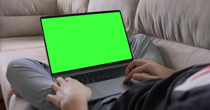 Man Uses MacBook Pro Laptop with Green Mock-up Screen While Sitting at the Sofa in His Cozy Living Room. 
