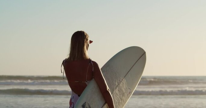 Young female looks out to ocean to gauge the conditiions to surf as waves roll onto shore in the distance