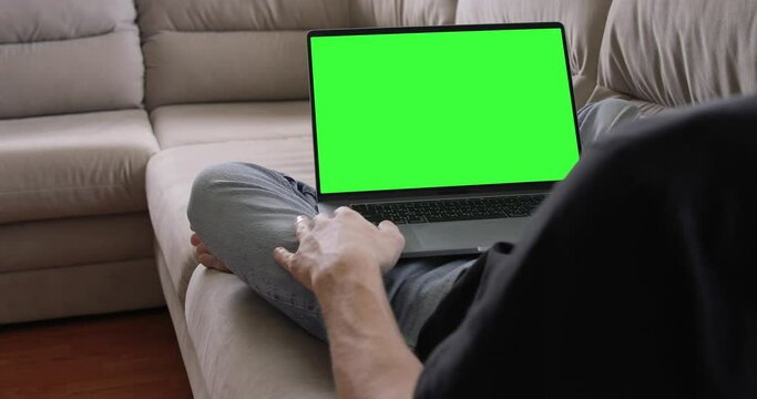 Man Looking on MacBook Pro Laptop with Green Mock-up Screen While Sitting at the Sofa in His Cozy Living Room. 