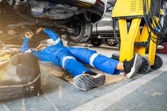 Mechanic Asian man underneath car fixing repairing front axle beam automobile vehicle parts examining using tools equipment working hard in workshop garage support and service in overall work uniform