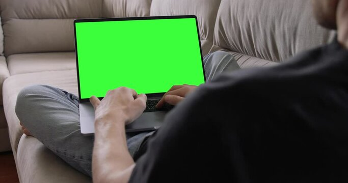 A man working on a MacBook Pro laptop with empty green screen monitor while lying in a lotus pose on the home sofa. Self-isolation, freelancing, remote work