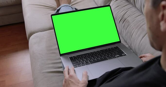 Man sitting on sofa in front of MacBook Pro laptop with green screen, chroma key, makes video call to friends, family, looking into display.