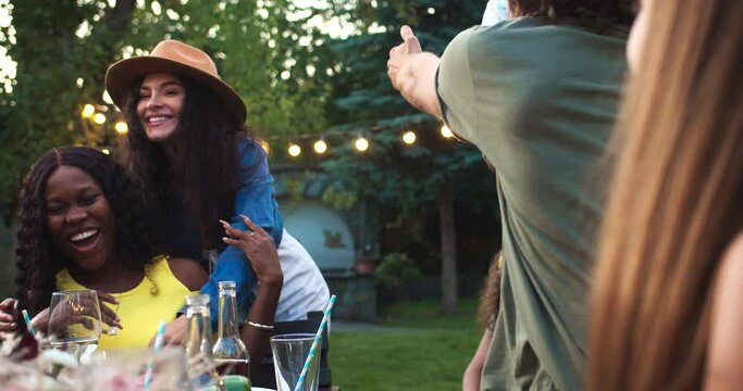 Multi-ethnic people sitting at table in garden. Caucasian female hugging African American girl. Cheerful guys and girls spend time together. Asian woman giving high five. Raising glasses Party concept