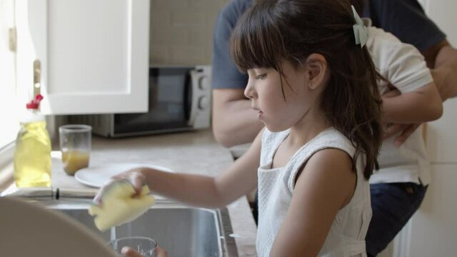 Girls rubbing glass with sponge and foam near flowing water. Dad teaching little son to wash dish. Side view. Family and household chores concept