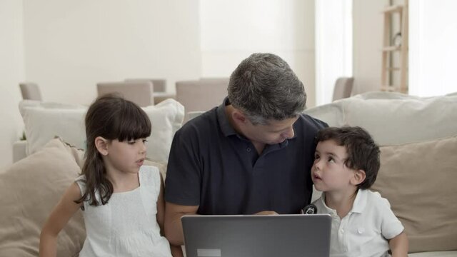Cheerful dad and two cute kids sitting on couch at laptop, watching movie, boy sneezing, father and sister touching his nose. Front view. Family leisure time concept