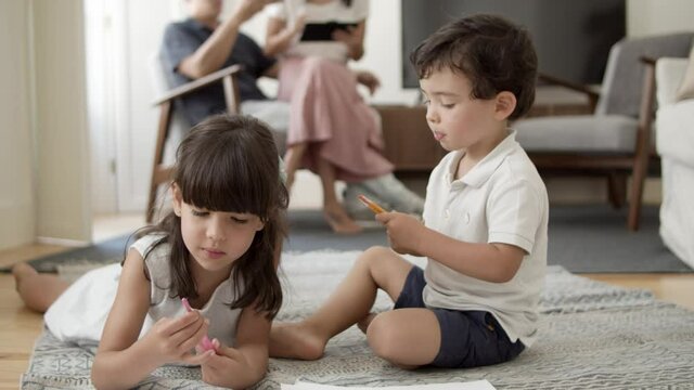 Sweet little boy and girl with markers drawing in living room while parents sitting together in background. Childhood or kids creative hobby concept