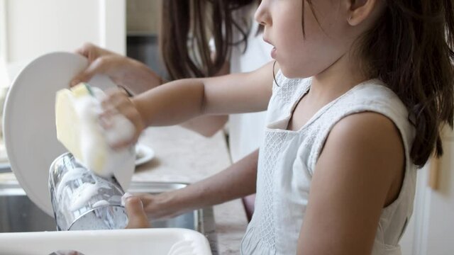 Focused girl helping her mom to wash dish, rubbing glass with sponge and soap foam. Side view, vertical motion. Family and household chores concept