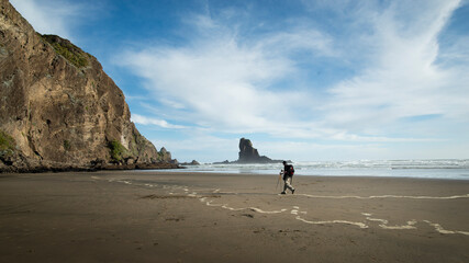 A backpacker walking on the black-sand Anawhata beach with keyhole rock in the distance, Waitakere, Auckland