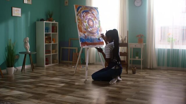 Young woman artist is working on a new masterpiece, standing on her knees to paint the bottom of the picture, applies acrylic dye with a wide brush, works with sincere pleasure, Slow motion.