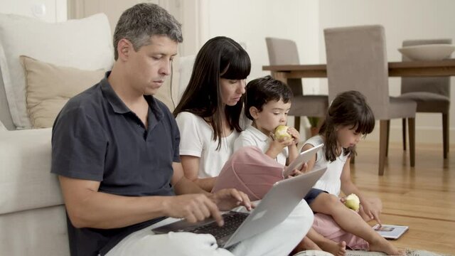 Focused parents couple using computers while children sitting near them and eating fresh apples in apartment. Communication and parenthood concept