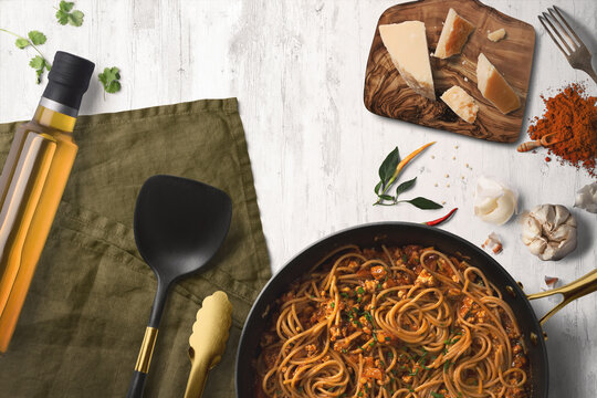 Top view kitchen scene showing a pan full of spaghetti bolognese surrounded by parmesan cheese of a chopping board, olive oil, a serving spoon and herbs and spices. 