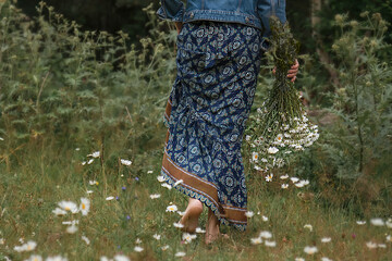 A girl walks barefoot through a clearing of daisies with a bouquet