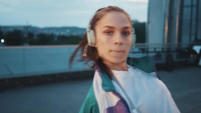 Portrait of stylish female dancer with headphone performing dance show looking at camera outdoors. Freestyle dancing improvisation. Brunette sporty woman listening to music moving by rhythm.