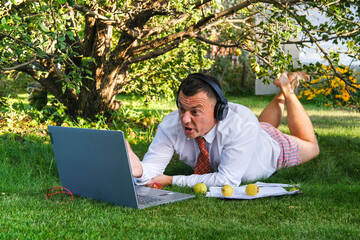 A male manager works remotely at home, holds an online meeting. He lies on the lawn in the garden. He talks emotionally with colleagues. He is wearing a shirt with a tie, underpants. Stay home and