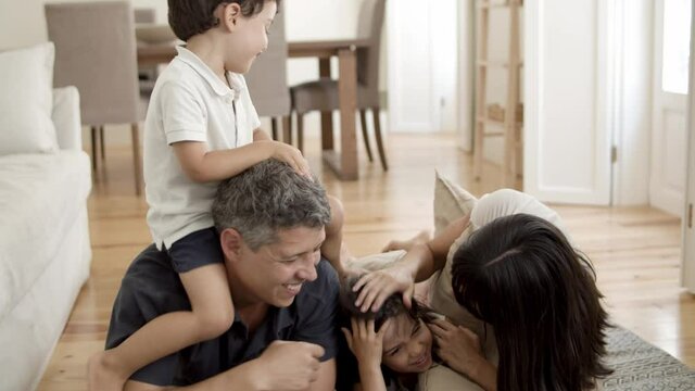 Joyful family with two kids relaxing at home, lying on carpet on floor in living room. Joyful little son sitting on dads neck while mom and kicking sister for fun. Family fun time concept