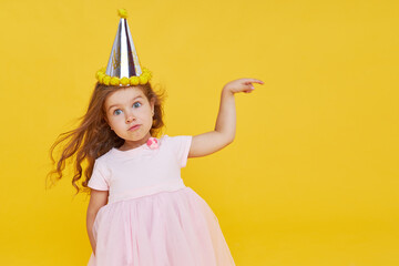 Party time. A joyful cute little child girl in a festive cap and elegant dress celebrates her birthday on a yellow background. Empty space for text. Pointing to the right