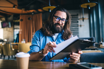 Smiling caucasian bearded 40 years man reading information from planner owning small business,...