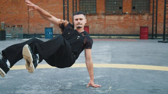 Male dancer showing break dance stunts on the roof. Professional artist dancing outside. Stylish attractive brunette man in black clothes dances looking at camera. Hip-hop freestyle battle. 4K