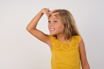 Young blonde kid girl wearing yellow dress over white background very happy and smiling looking far away with hand over head. Searching concept.