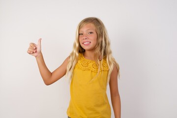 Young blonde kid girl wearing yellow dress over white background. Looking proud, smiling doing thumbs up gesture to the side. Good job!