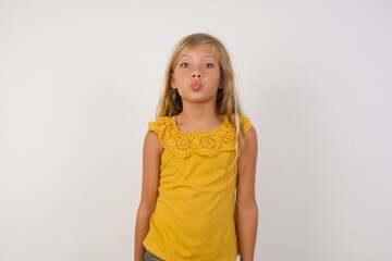 Shot of pleasant looking Young blonde kid girl wearing yellow dress over white background pouts lips, looks with green eyes at camera,  has fun with girlfriend, Human facial expressions