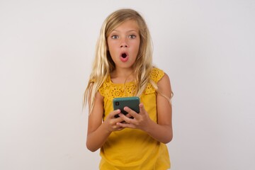 Shocked Little girl with beautiful blonde hair over white background. opens mouth hold phone reading advert unbelievable big shopping prices