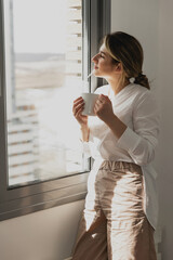 Young beautiful woman near window with cup of coffee or tea at home. Dream and relax. Good day. Woman in the morning is holding a mug and looking at the sunrise and having a perfect cozy morning