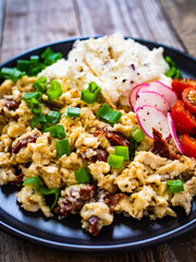 Delicious breakfast - scrambled eggs with fried sausages, cottage cheese and vegetables served on black plate on wooden table
