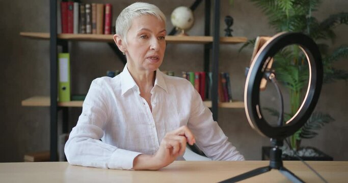 Elderly woman teacher in white shirt doing online lesson using smartphone with LED ring light