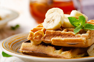 Belgian waffles served with banana and mint leaf on white wooden kitchen table with syrup aside