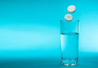 Transparent glass with clean water and pills flying into a glass on a blue background close-up. Dissolving medicine drug, antibiotic or vitamin. Copy space. 