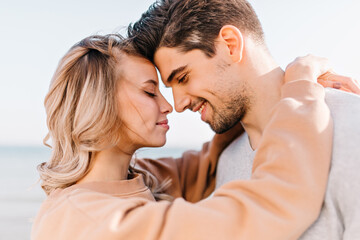 Fototapeta premium Elegant girl embracing husband with eyes closed. Outdoor photo of romantic couple posing on the street.