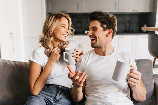 Cheerful young man telling his girlfriend funny joke and making her smiling. Attractive couple at home drinking coffee looking at each other and laughing.