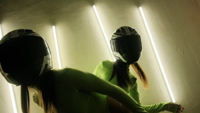 Two girls in motorcycle helmets are dancing in a nightclub. A pj with nice bodies and light green bodysuits. Neon light on the wall.
