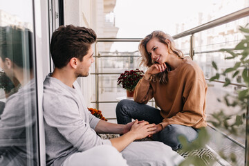 Curious caucasian girl talking with friend at terrace. Dreamy young lady sitting at balcony with boyfriend. - obrazy, fototapety, plakaty