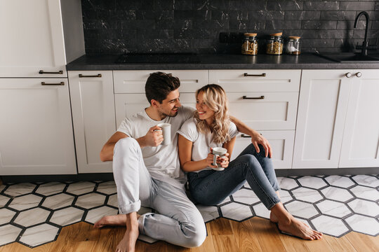 Blissful couple drinking coffee in stylish kitchen. Indoor photo of romantic girl looking at boyfriend while enjoying tea.